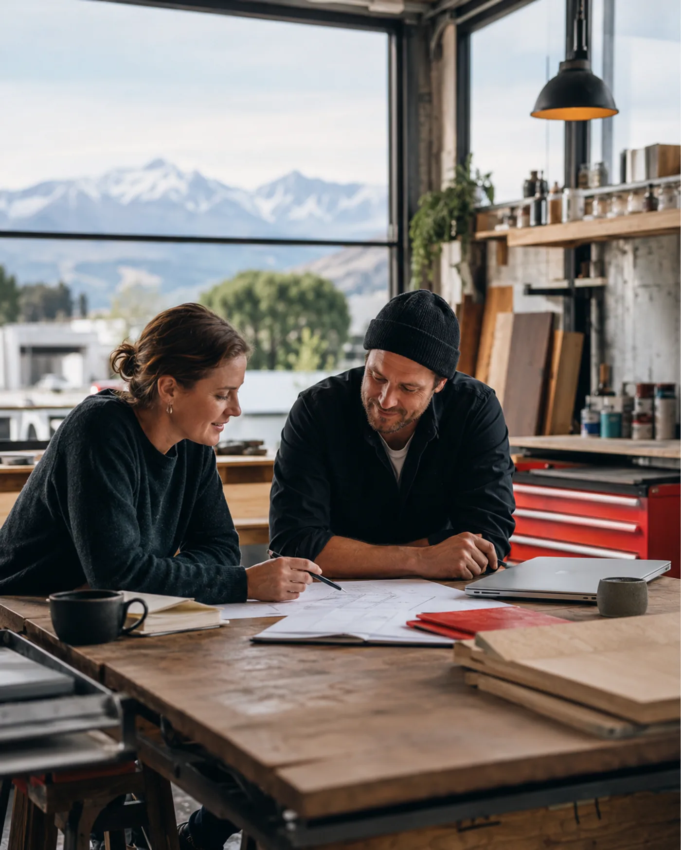 Two Canterbury business owners reviewing plans together in a workshop, the Southern Alps through the window